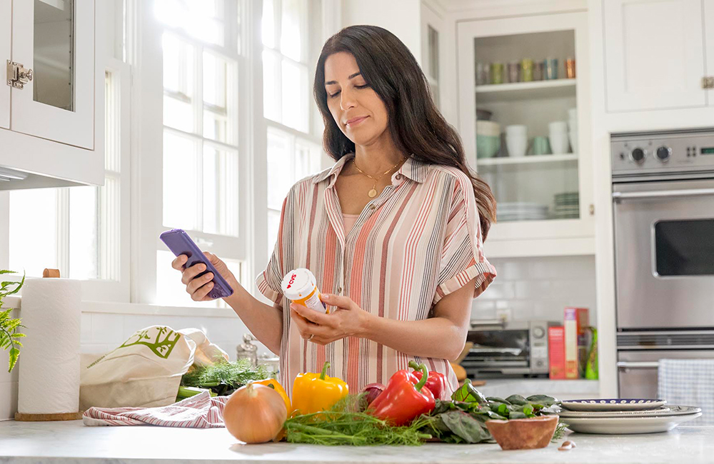 A woman stands in her kitchen with her groceries to examine her prescription bottle.