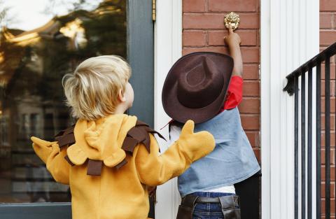 A child in a cowboy costume rings a doorbell while trick-or-treating with another child, who is dressed as a lion.